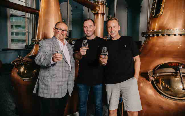 Three men with whiskey glasses in front of copper stills