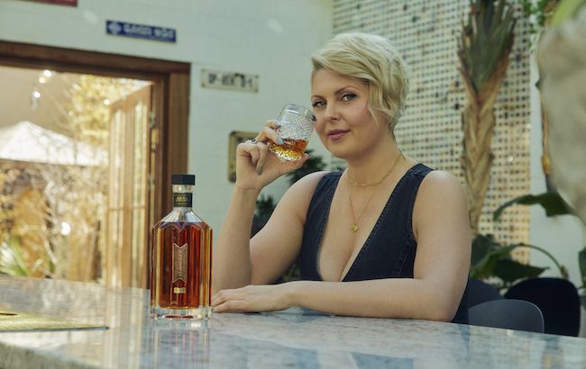 A woman sits at a counter top with a glass of whiskey