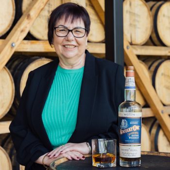 A woman in front of whiskey barrels with a bottle of bourbon