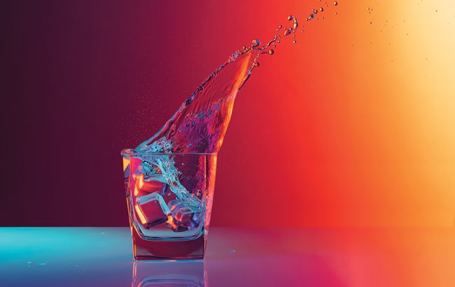 Taste Masters - Ice cube falls into a transparent glass of water standing on mirror surface over gradient red orange background in neon light. Art, beauty, drinks
