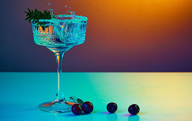 Portrait with tasty and fresh vodka cocktail in glass with splashes and cherryes over gradient background in neon glow. Party club entertainment. Mixed light. Concept of celebration, bar, ad