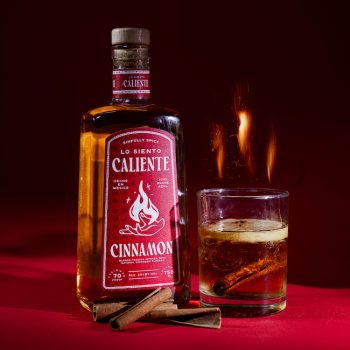 A bottle of cinnamon infused tequila with a glass emitting heat and cinnamon sticks next to it
