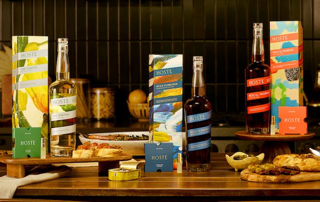 Three Hoste bottled cocktails are shown on a table surrounded by food