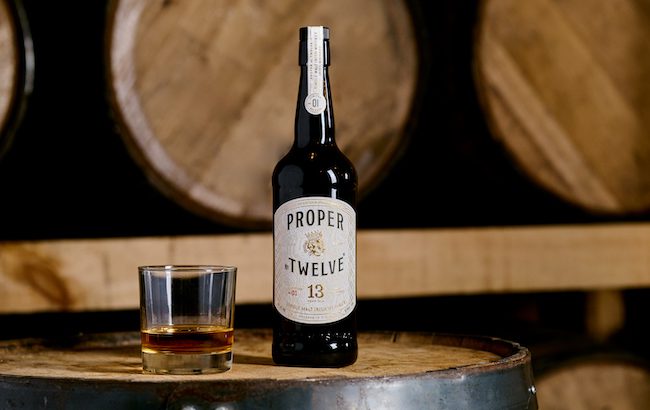 A bottle of Proper Twelve 13 year old single malt on a barrel with a whiskey glass next to it