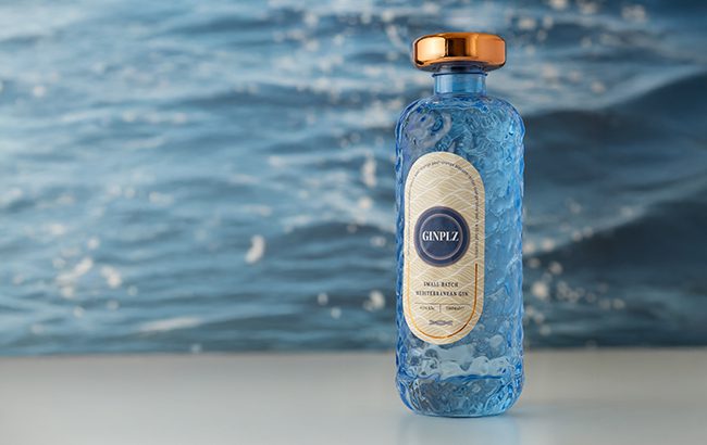 3_GINPLZ bottle with golden cap set before rippling blue water, evoking Mediterranean freshness