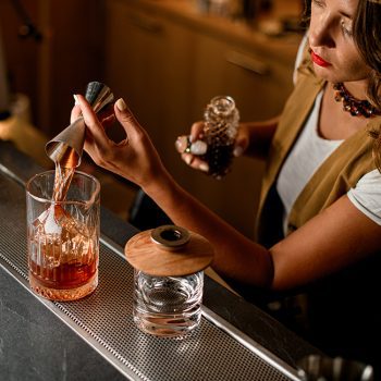 Top view of bar counter with mixing glass in which female bartender pours brown drink from jigger