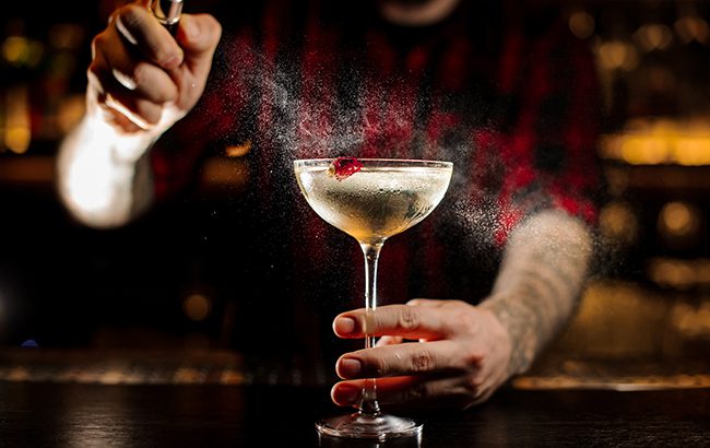 Bartender spraying on a glass of a delicious Twinkle cocktail with one berry on the bar counter on the blurred dark and yellow light background