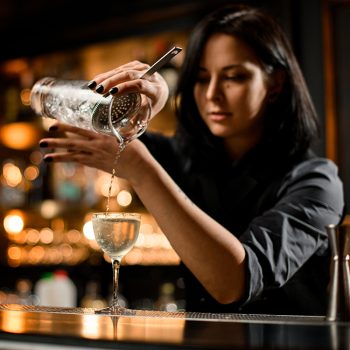 global bar report - Professional bartender girl pouring a trasparent alcoholic drink from the measuring cup to the glass through the strainer filter on the bar counter