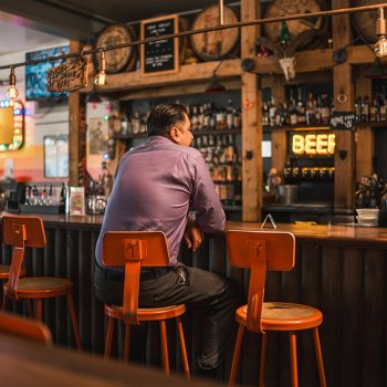 A male with Hispanic features enjoys solitude with a drink, seated casually at a cozy bar, surrounded by an array of bottles and bar stools.